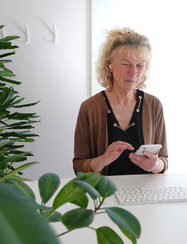Susanne sitter vid ett skrivbord, håller mobilen i handen och har airopds i öronen