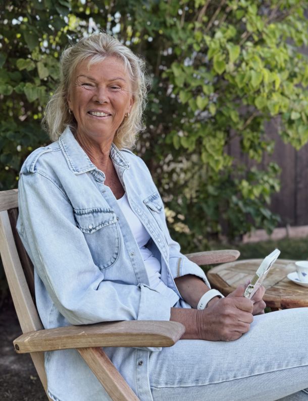 Susanne sitter ute med gröna buskar bakom sig och med en mobil i handen.
